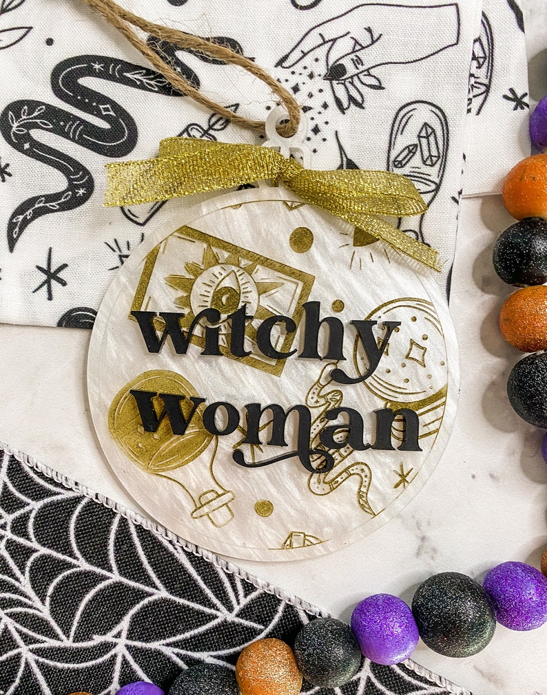May include: A white round ornament with a gold glitter finish and black lettering that reads "Witchy Woman". The ornament has a gold ribbon and a twine hanger.