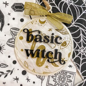 May include: A white round ornament with gold glitter and black lettering that says "basic witch". The ornament has a gold ribbon and is hanging from a string.