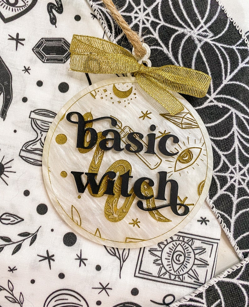 May include: A white round ornament with gold glitter and black lettering that says "basic witch". The ornament has a gold ribbon and is hanging from a string.