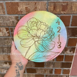 K&ouml;nnte beinhalten: Ein rundes Holzschild mit einem Regenbogen-Ombre-Hintergrund und einem goldenen Blumendesign. Das Jahr 2023 ist in Gold auf der rechten Seite des Schildes eingraviert.