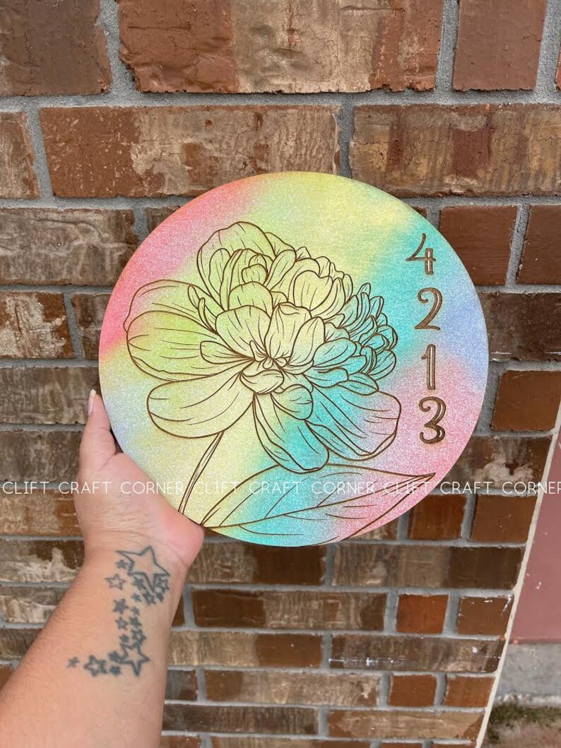 K&ouml;nnte beinhalten: Ein rundes Holzschild mit einem Regenbogen-Ombre-Hintergrund und einem goldenen Blumendesign. Das Jahr 2023 ist in Gold auf der rechten Seite des Schildes eingraviert.