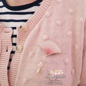 Könnte beinhalten: Eine rosafarbene und weiße Perlenbrosche in Fächerform mit einer goldenen Nadel. Die Brosche ist an einem rosafarbenen Strickcardigan befestigt.