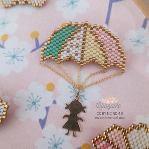 May include: A beaded pendant necklace with a rainbow design, gold accents, and a gold girl silhouette. The necklace is set against a pink floral backdrop.
