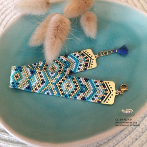 Peut inclure: Un bracelet en perles bleu, blanc et or avec un motif géométrique. Le bracelet a un fermoir en or et un pompon bleu.