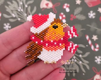 Puede incluir: Un broche navideño de petirrojo hecho con cuentas. El pájaro lleva un gorro y una bufanda rojos y blancos. El cuerpo del petirrojo es naranja, marrón y blanco. El broche está hecho de pequeñas cuentas de colores.