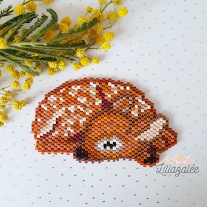 Puede incluir: Una broche de animal hecha con cuentas en forma de erizo enroscado. El erizo está hecho con cuentas marrones, naranjas, blancas y negras. La broche está sobre un fondo blanco con purpurina dorada.