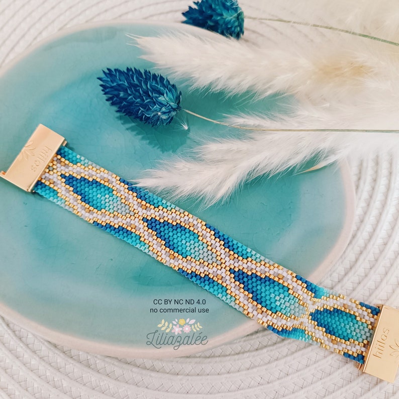May include: A beaded bracelet with a geometric pattern in shades of blue, white, and gold. The bracelet has gold metal clasps on each end. The bracelet is on a blue and white surface.