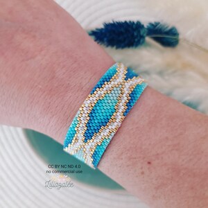 May include: A blue, white, and gold beaded bracelet with a geometric pattern. The bracelet is on a person's wrist.