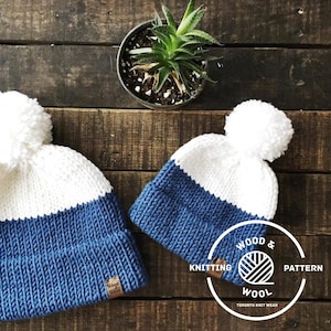 Op de afbeelding: Twee gebreide mutsen met witte pompoenen. De grotere muts is blauw en wit met een witte band. De kleinere muts is blauw en wit met een witte band. De mutsen liggen op een houten oppervlak. De tekst "Wood & Wool Knitting Pattern" staat op een bruin label.