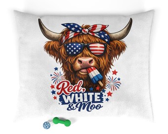 Op de afbeelding: Een wit kussen met een afbeelding van een Schotse hooglander met een zonnebril en een patriottische hoofdband. De koe houdt een ijsje vast en de tekst luidt "Red, White & Moo". Vuurwerk en sterren omringen de koe.