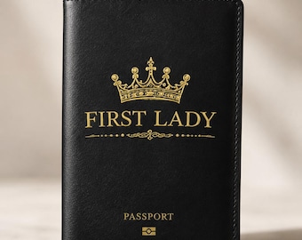 Op de afbeelding: Zwarte paspoorthouder met gouden tekst en een kroonontwerp. De woorden "FIRST LADY" staan boven de decoratieve lijn, met "PASSPORT" eronder. Onderaan staat een klein paspoortpictogram.