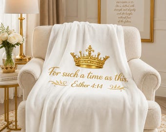 Op de afbeelding: Een witte deken met een gouden kroonontwerp en de tekst "For such a time as this. Esther 4:14" in gouden letters. De deken ligt over een crèmekleurige fauteuil. Een decoratief item voor in huis.