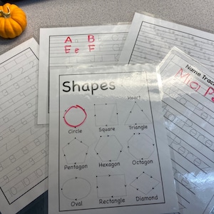 May include: Educational worksheets for tracing numbers, letters, and shapes. The "Shapes" worksheet features a circle, square, triangle, and other geometric figures. A small orange pumpkin sits on the top left corner.
