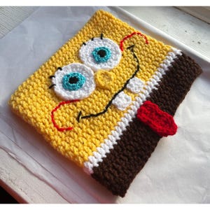 May include: A handmade yellow crochet item resembling a cartoon character's face. The item features large white and blue eyes, a black stitched mouth, and a red tongue. The edges are brown, white, and yellow.