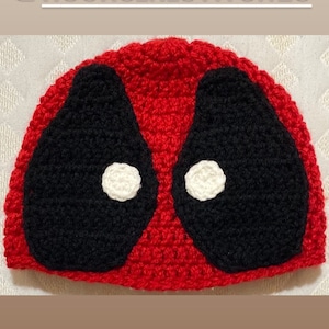 May include: A red and black crocheted hat with a design featuring large black eye shapes and small white circles. The hat is handmade and features the text "@HOOKS_N_STITCHES".