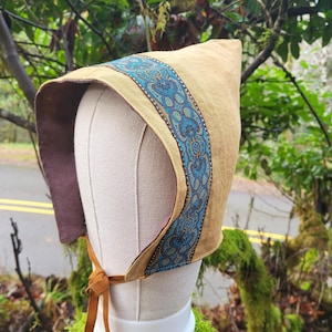 May include: A brown linen bonnet with a blue and gold patterned trim. The bonnet has a long brown tie.