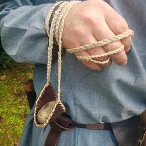 May include: A handmade leather and woven cord sling, with a stone inside. The sling is attached to a braided cord wrapped around a hand. The sling is brown with a tan braided cord. The person is wearing a blue shirt.