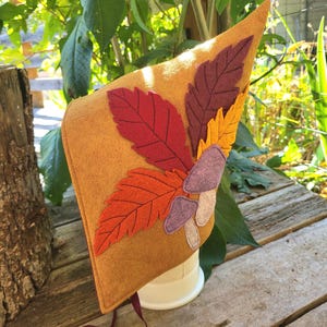 May include: A mustard yellow felt bonnet featuring an appliqué of three felt leaves in red, dark purple, and orange, along with two small felt mushrooms in light purple and beige. The bonnet has a pointed shape and includes thin dark red ties.