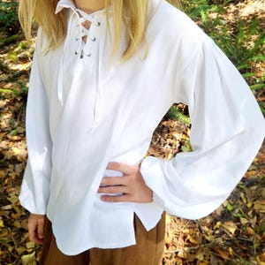 May include: White long-sleeved shirt with a lace-up neckline and a split hem. The shirt has puffy sleeves and is made of a lightweight fabric. The shirt is paired with brown pants.