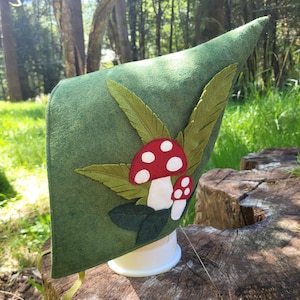 May include: A green felt hat with a pointed tip, decorated with felt leaves and red mushrooms with white spots. The hat is displayed on a white stand, set against a backdrop of trees and greenery.