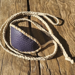 May include: A handmade slingshot with a dark purple leather pouch and a braided natural rope. The pouch is stitched with a light tan thread. The slingshot is lying on a weathered wooden surface.