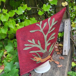 May include: A deep red pointed bonnet featuring an appliqued light green leafy branch design and a brown snail at the base. The bonnet is constructed from a textured fabric and includes thin red fabric ties hanging from the bottom edge.