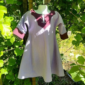 May include: A light purple tunic with three-quarter length sleeves and a dark maroon V-shaped neckline panel. The garment features a central placket with four small white buttons and matching dark maroon cuffs on the sleeves, displayed on a mannequin.