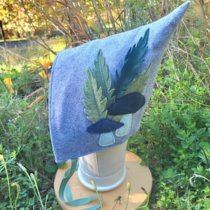 May include: A light blue felt bonnet featuring a pointed crown and long ribbon ties. The side is decorated with layered fabric appliques of green fern leaves and two dark blue mushrooms with light blue stems, creating a textured botanical design.