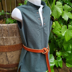 May include: A sleeveless dark teal tunic featuring a stand-up collar and a front placket with four metal eyelets. The garment is cinched at the waist with a tan leather belt that features a round metal buckle and a long hanging strap end.