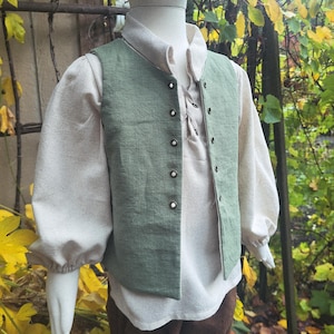 May include: A child's costume featuring a light green vest with dark buttons over a cream-colored shirt with a lace-up neckline. The sleeves are long and gathered at the wrists. The costume is completed with brown pants.