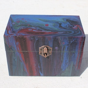 Puede incluir: Una caja de madera con un diseño de pintura marmoleada azul, verde y roja. La caja tiene un cierre de latón.