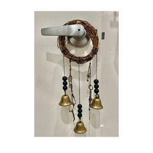 May include: A brown twig wreath with black beads and gold keys hanging from it. The wreath is attached to a door with a silver door handle. There are three gold bells hanging from the wreath.