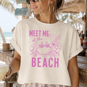 Könnte beinhalten: Cremefarbenes Crop-Top mit dem Text "MEET ME at the BEACH" in Pink, mit einer Krabbe und einem Cocktail-Motiv. Die Person trägt eine Sonnenbrille und eine gestreifte Hose und hält eine Margarita. Die Szene spielt in einer Strandbar.