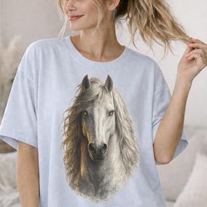 Könnte beinhalten: Hellblaues T-Shirt mit einer detaillierten Illustration eines weißen Pferdekopfes und einer fließenden Mähne. Das Pferd hat eine hellbraune Mähne und ist auf dem T-Shirt zentriert. Das Model trägt eine rosa-weiß karierte Shorts.