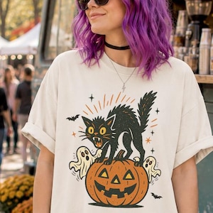 Könnte beinhalten: Ein cremefarbenes T-Shirt mit einem Grafikdruck einer schwarzen Katze mit gekrümmtem Rücken, die auf einem geschnitzten orangefarbenen Kürbis steht, umgeben von zwei kleinen weißen Geistern, Fledermäusen und Sternen, kombiniert mit einer schwarz-grau gestreiften Hose.