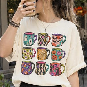May include: A cream-coloured short-sleeve t-shirt featuring a graphic print of nine distinct mugs arranged in a grid. Each mug displays a different colourful pattern, including ghosts, pumpkins, black cats, spider webs, and candy corn motifs.