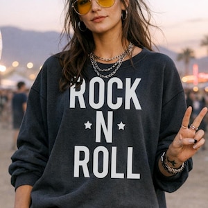 May include: A dark gray sweatshirt with the text "ROCK *N* ROLL" in white. The person is wearing denim shorts, sunglasses, and jewelry. A Ferris wheel and crowd are in the background.
