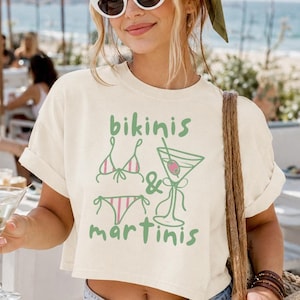 Könnte beinhalten: Cremefarbenes Crop-Top mit den grünen Worten "bikinis & martinis". Das Shirt wird mit Jeansshorts und einer weißen Sonnenbrille kombiniert. Eine Strohtasche hängt über der Schulter. Die Szene spielt sich im Freien in Strandnähe ab.