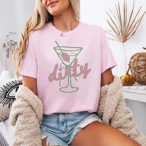 May include: A light pink t-shirt featuring a green and pink martini glass design with the word "dirty". The graphic includes an olive and a straw. The shirt is a casual, everyday wear item.