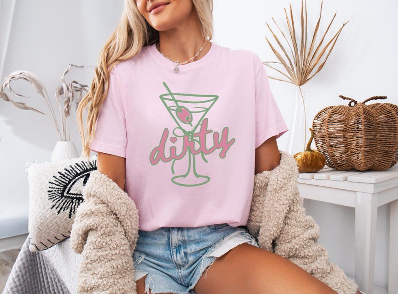 May include: A light pink t-shirt featuring a green and pink martini glass design with the word "dirty". The graphic includes an olive and a straw. The shirt is a casual, everyday wear item.