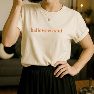 May include: Cream-colored t-shirt with the text "halloween slut." A black cat-shaped mug is held up. The person is wearing a black skirt and a silver necklace.