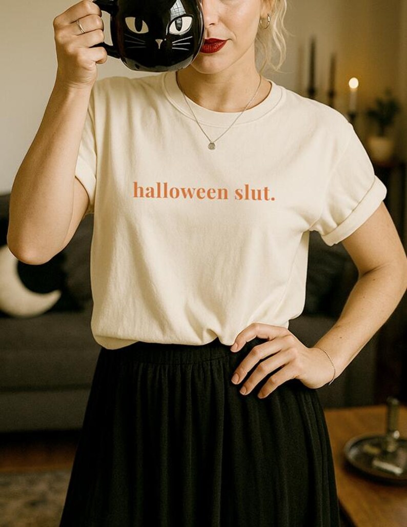 May include: Cream-colored t-shirt with the text "halloween slut." A black cat-shaped mug is held up. The person is wearing a black skirt and a silver necklace.