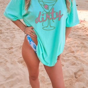 May include: A light turquoise t-shirt with a cocktail graphic and the word "dirty" in pink script. The shirt is worn with a floral print bikini bottom. The image is taken on a sandy beach.