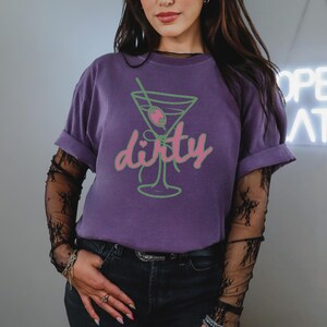 May include: A purple t-shirt with a green and pink martini glass graphic and the word "dirty". The shirt has rolled-up sleeves and is paired with black jeans. The person is wearing a black lace long sleeve shirt underneath.