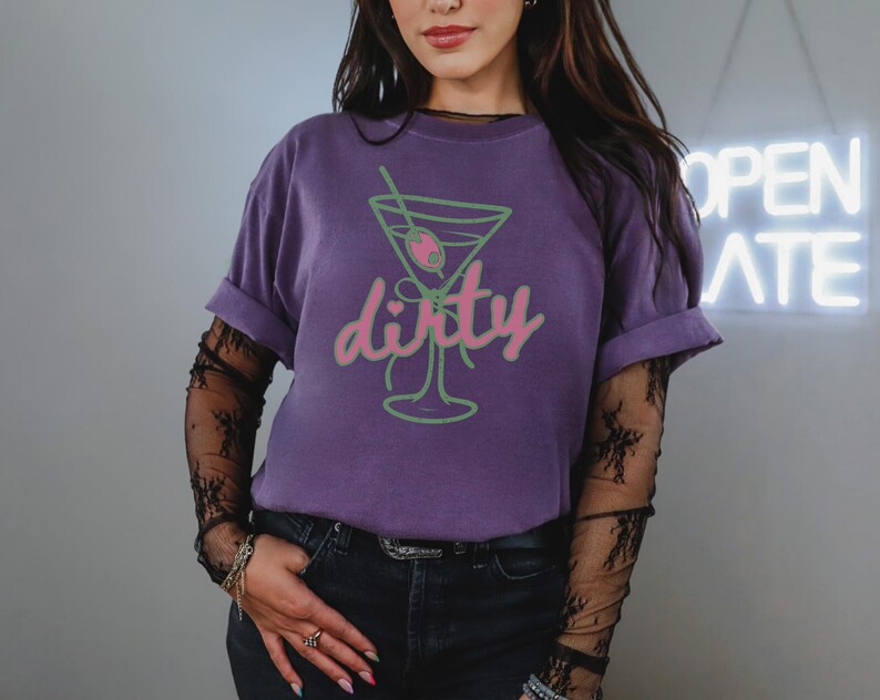 May include: A purple t-shirt with a green and pink martini glass graphic and the word "dirty". The shirt has rolled-up sleeves and is paired with black jeans. The person is wearing a black lace long sleeve shirt underneath.