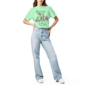 May include: A light green t-shirt featuring a martini glass graphic with the word "dirty" in a playful font. The shirt is paired with light blue jeans and white sneakers. The overall style is casual and relaxed.