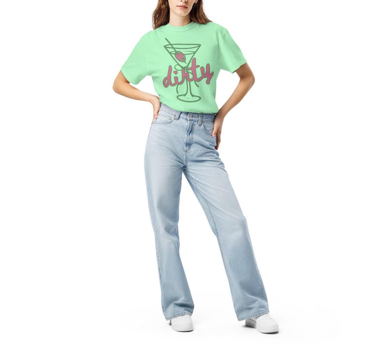 May include: A light green t-shirt featuring a martini glass graphic with the word "dirty" in a playful font. The shirt is paired with light blue jeans and white sneakers. The overall style is casual and relaxed.