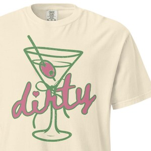 May include: Cream-colored t-shirt featuring a green martini glass with a pink olive and the word "dirty" in pink script. The shirt has a classic crew neck and short sleeves, perfect for casual wear.