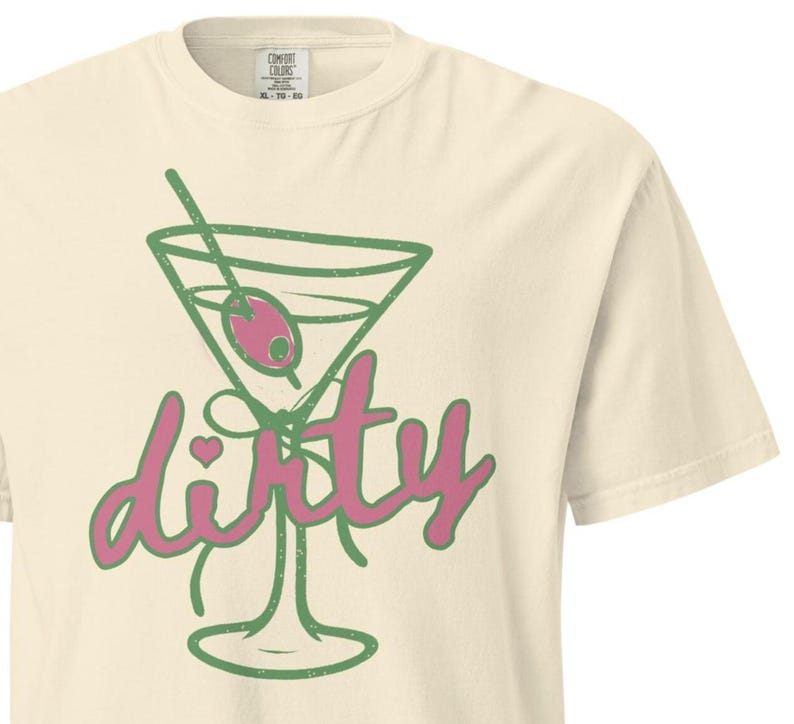 May include: Cream-colored t-shirt featuring a green martini glass with a pink olive and the word "dirty" in pink script. The shirt has a classic crew neck and short sleeves, perfect for casual wear.