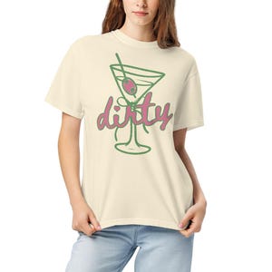 May include: Cream-colored t-shirt featuring a green outline of a martini glass with an olive and a straw. The word "dirty" is written in pink script below the glass. The shirt is made of a soft material.